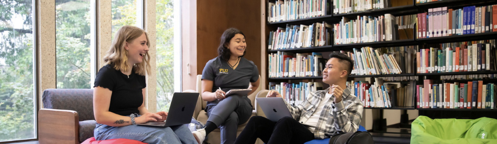 Three students in library in front of window and stack of books